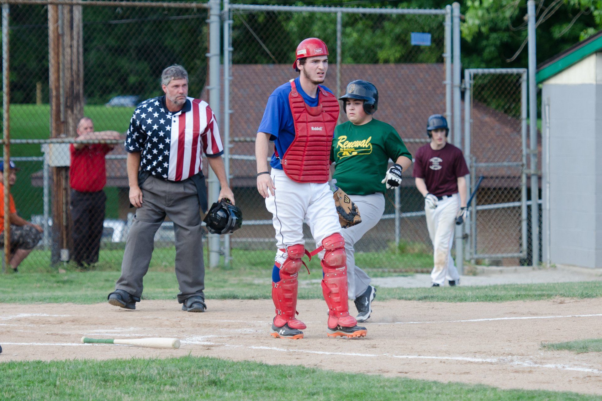 Baseball game: Catcher, batter, umpire, and runner on the field. The umpire wears an American flag shirt.