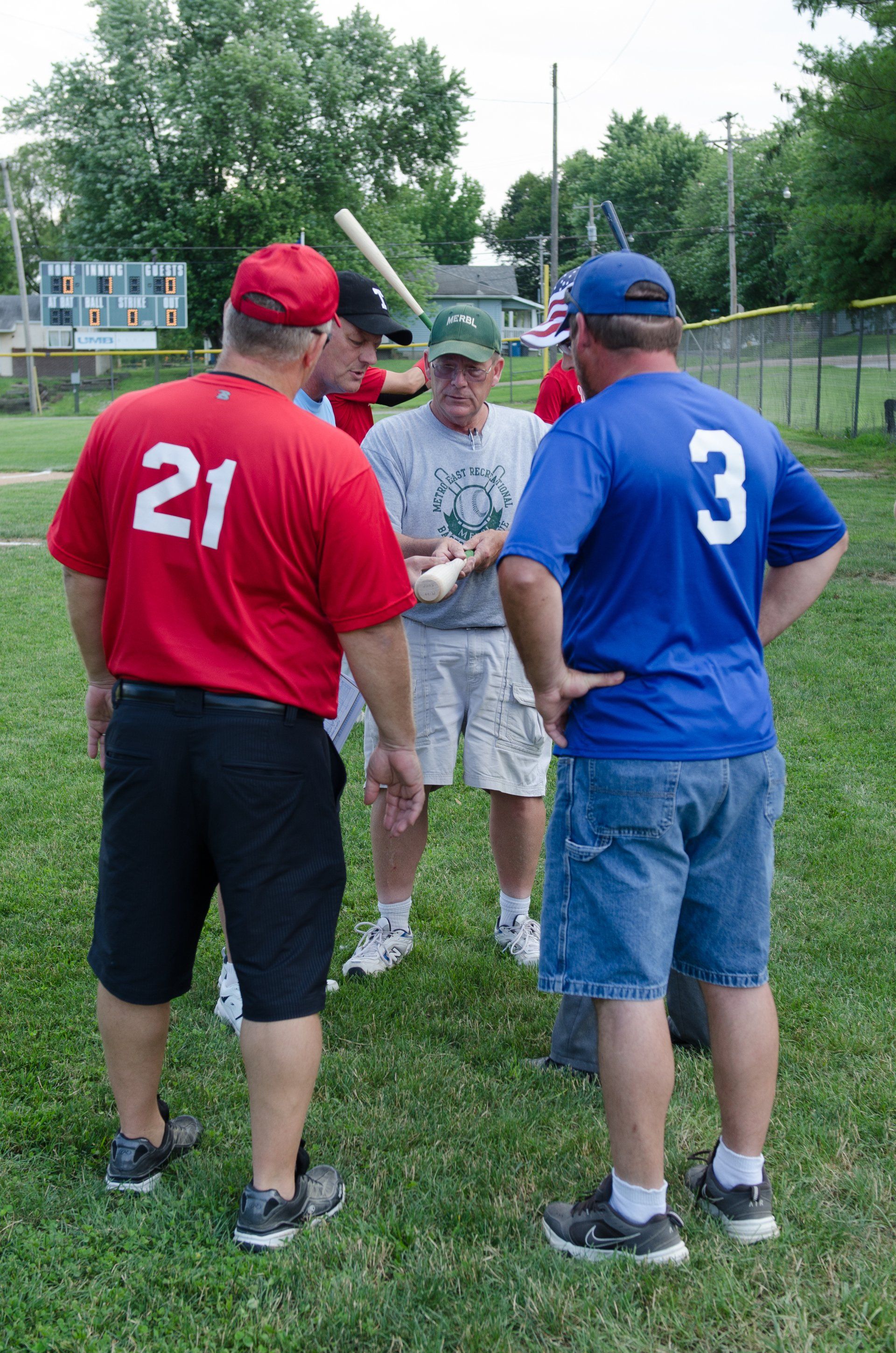 Baseball coaches huddle on a field. Men in red, blue, and green shirts converse; one holds a baseball.