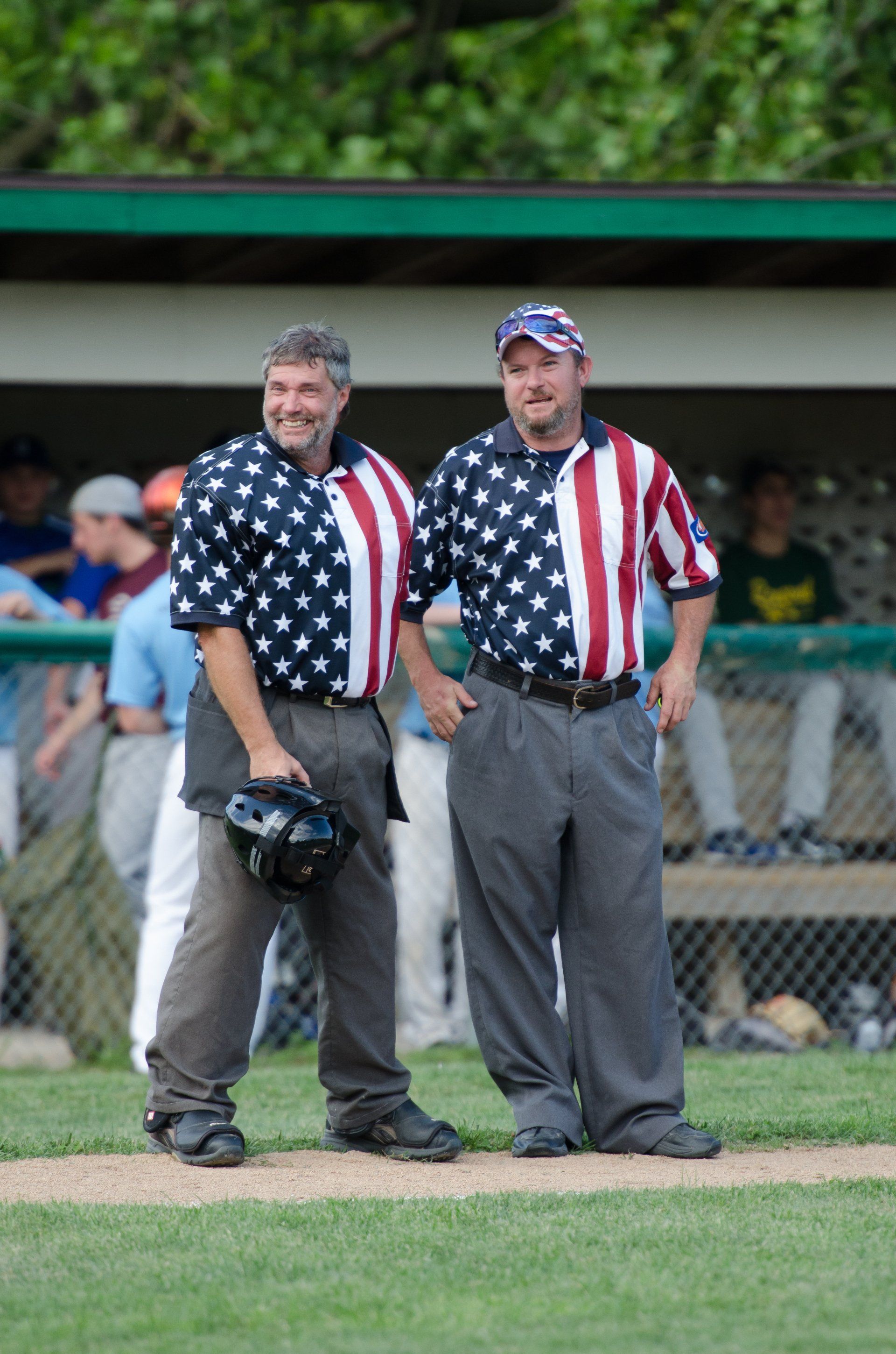 Two baseball umpires in patriotic shirts stand on a baseball field.