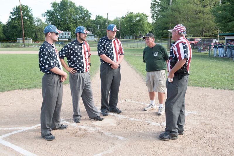 Baseball umpires in American flag shirts and hats consult with a man at home plate.