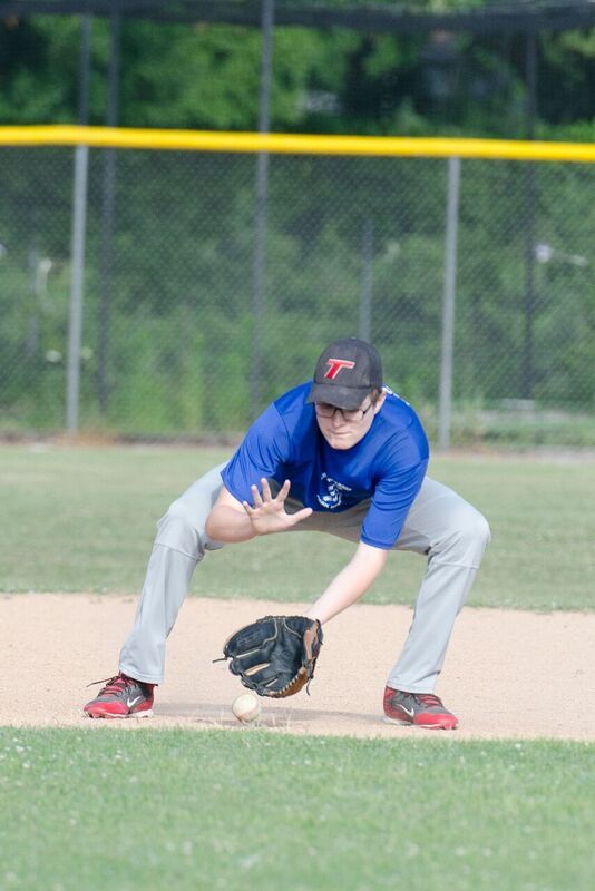 Baseball player fielding a ground ball, blue shirt, gray pants, and red shoes, on a baseball field.