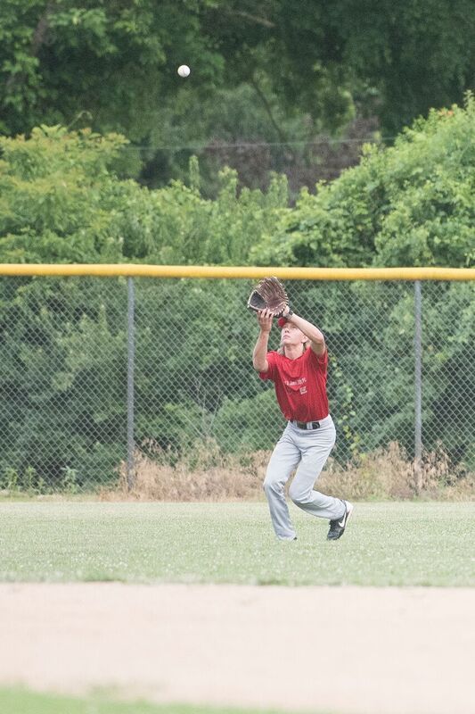 Baseball player jumps to catch a high fly ball, wearing a red shirt and gray pants on a baseball field.