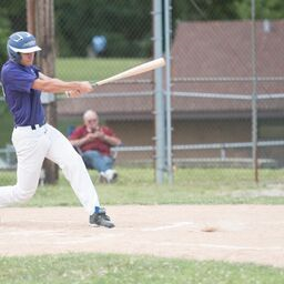 Baseball player swinging bat, wearing purple shirt, white pants, helmet; on a baseball field.