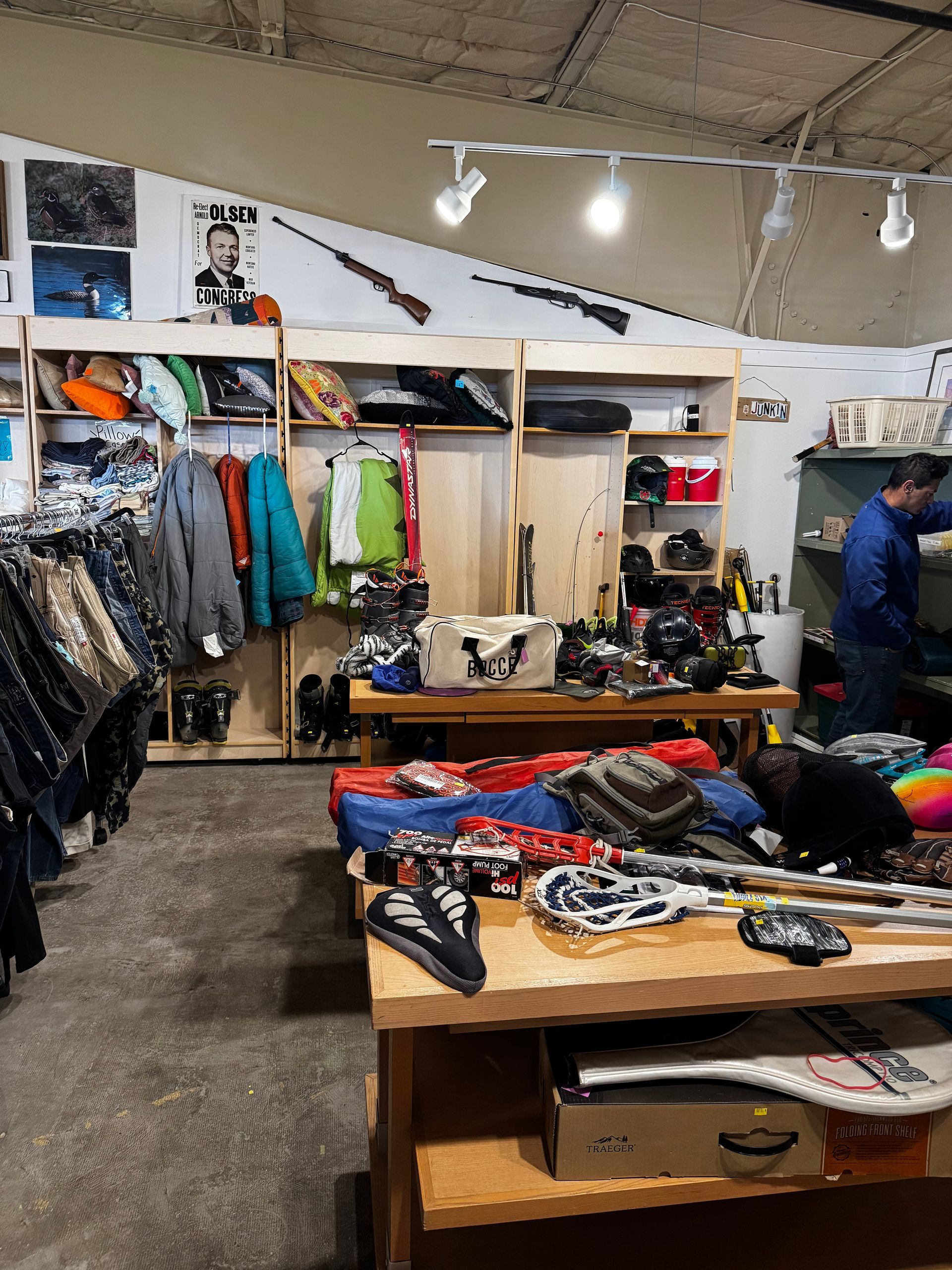 Inside a store selling used outdoor gear, items on shelves and tables, a person browses.
