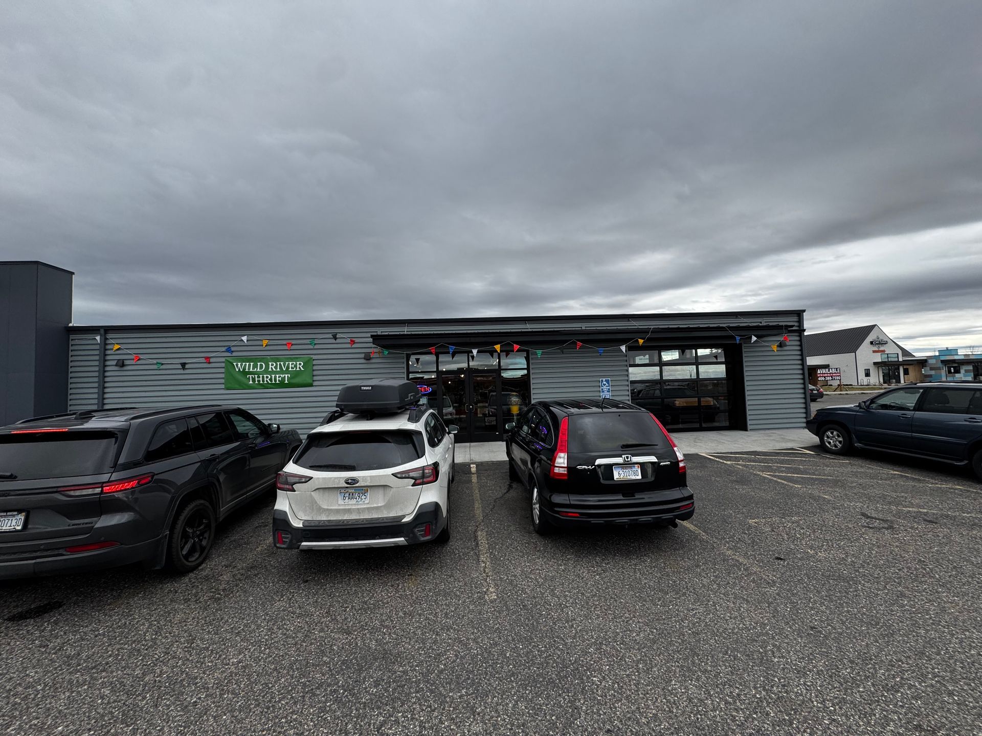 Exterior of a business with parked cars; gray building with green sign, cloudy sky.