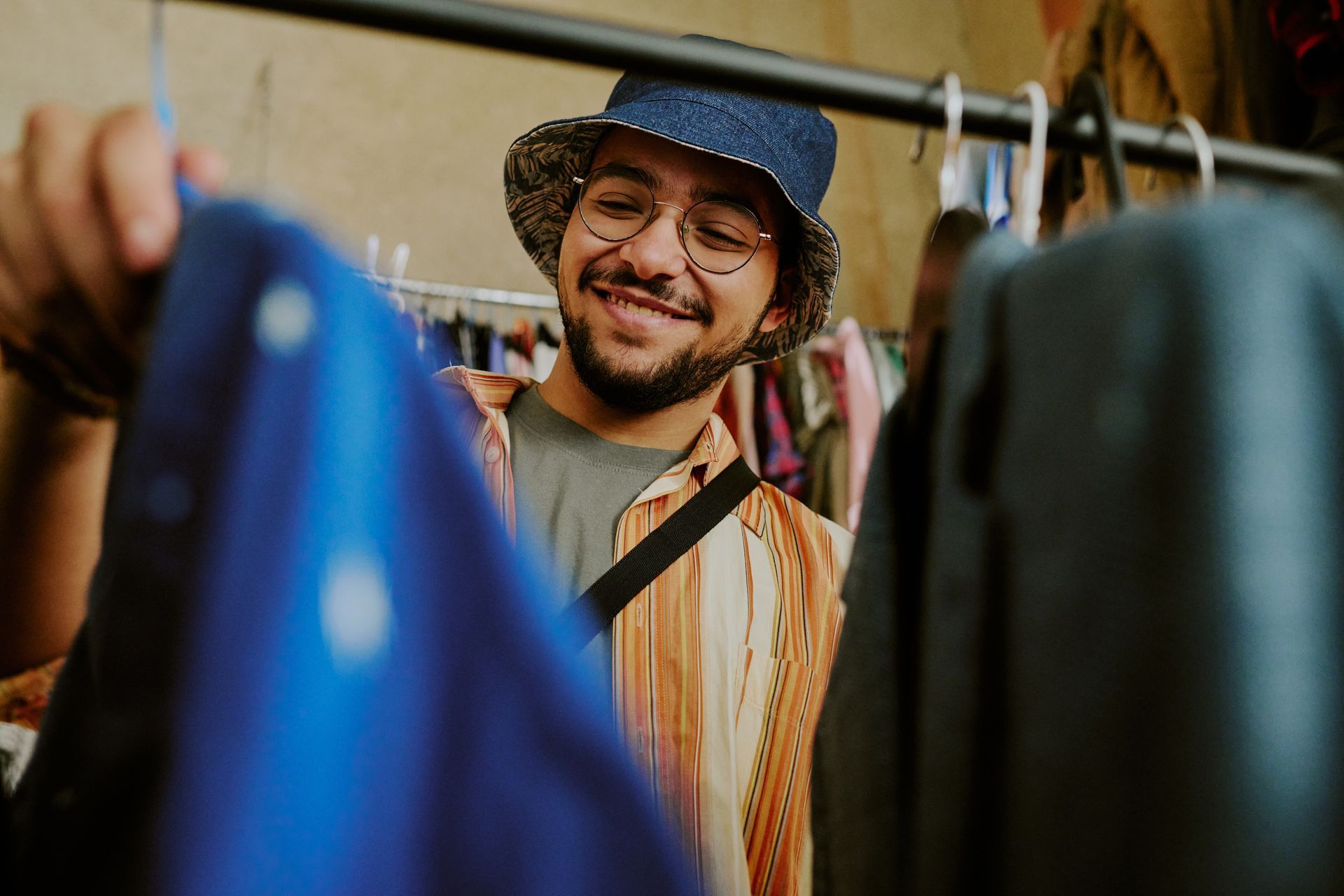 Smiling person with glasses and a bucket hat looking at clothes on a rack in a store.