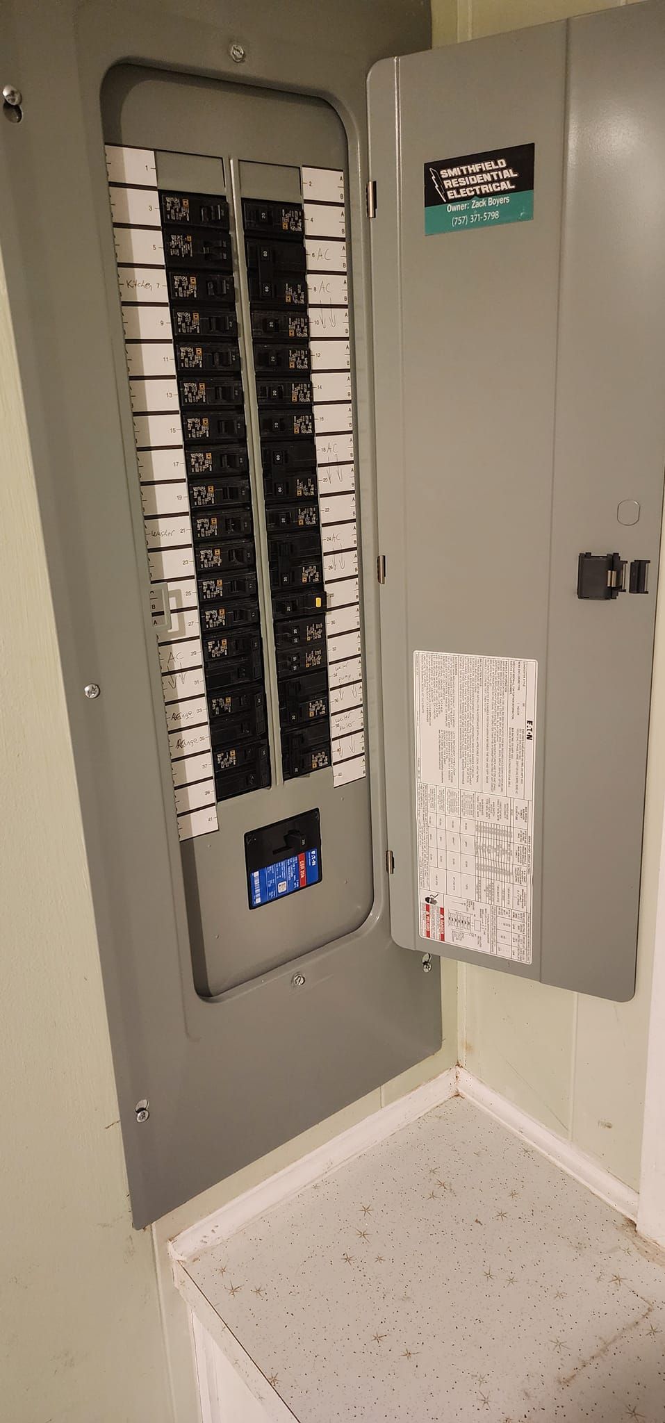 Open electrical panel in a corner. The door is ajar, revealing circuit breakers. The panel is gray, set in a light-colored room.