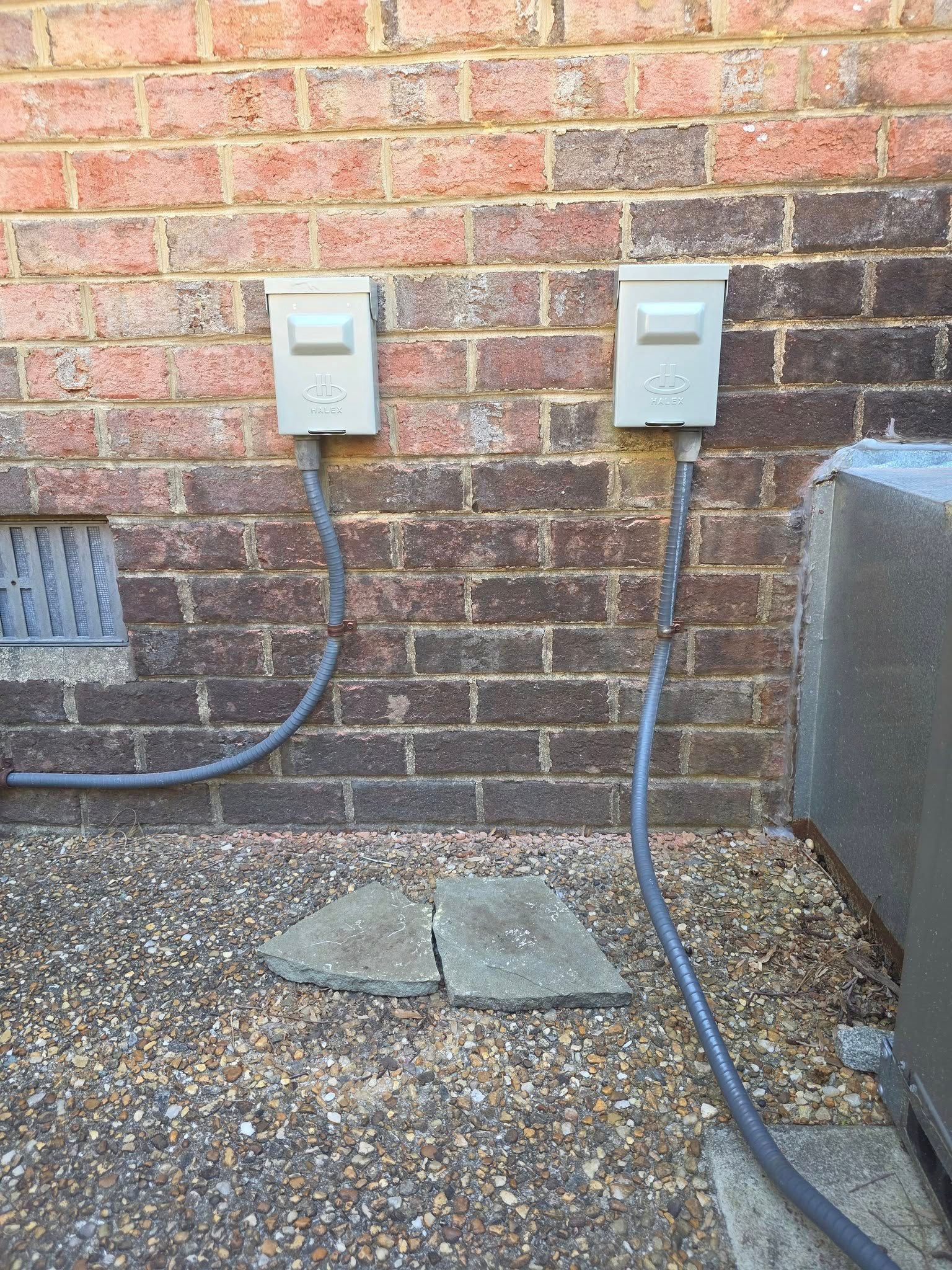 Two electrical boxes mounted on a brick wall with conduit running to the ground. Pebbles and two broken stones are in front of the wall.