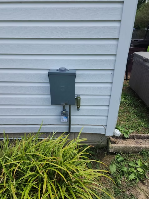 Outdoor electrical box mounted on light blue siding with a grounding rod and a faucet nearby. Yellow-green plants are in the foreground.