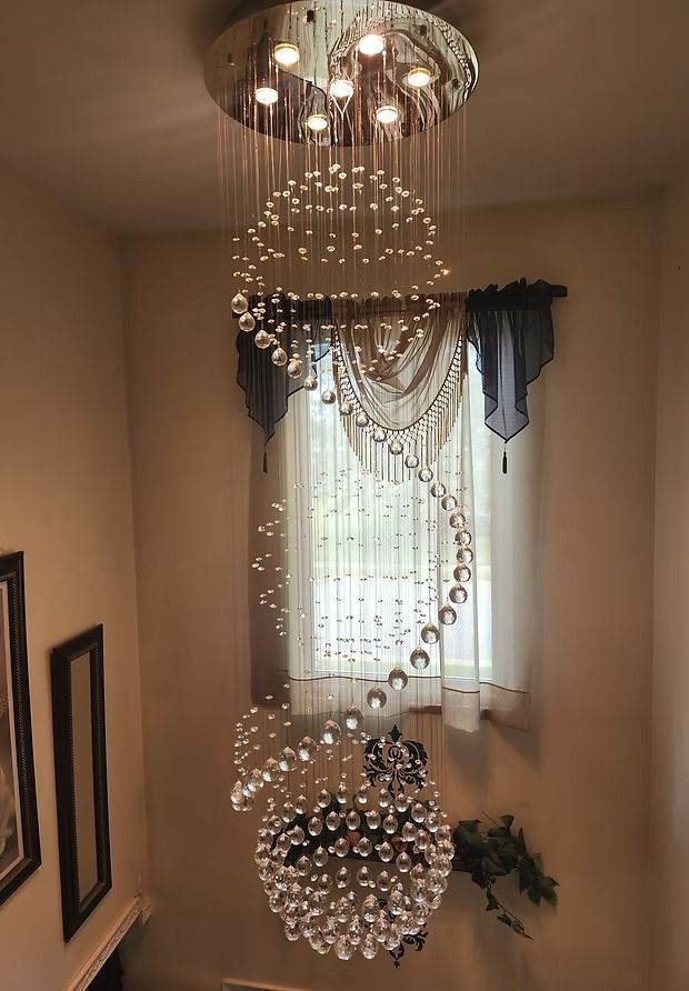 A modern, spiral-shaped chandelier with crystal-like orbs hanging in a stairwell, lit by overhead lights.