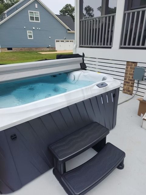 A hot tub on a deck with steps. It's gray with the top open, filled with blue water. A house is visible in the background.