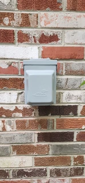 A gray electrical box is mounted on a brick wall with various colors like red, brown, and white.