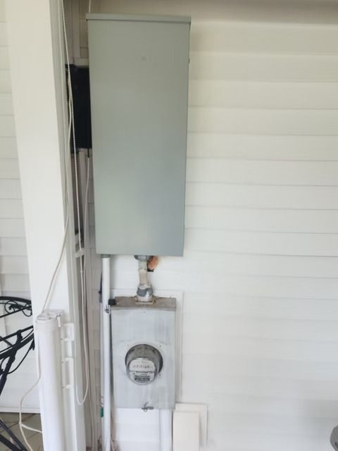 Electric meter box and service panel mounted on a white building exterior. A grey metal box sits above the meter.