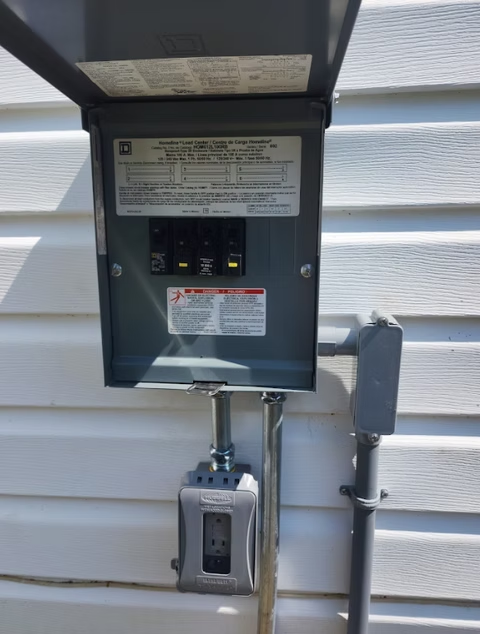 Exterior electrical box mounted on white siding, with the cover open to reveal circuit breakers. Conduit runs to a power outlet.