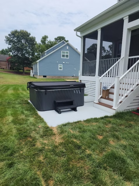 A hot tub sits on a concrete patio next to a screened porch. The scene is outdoors with grass and a blue house in the background.