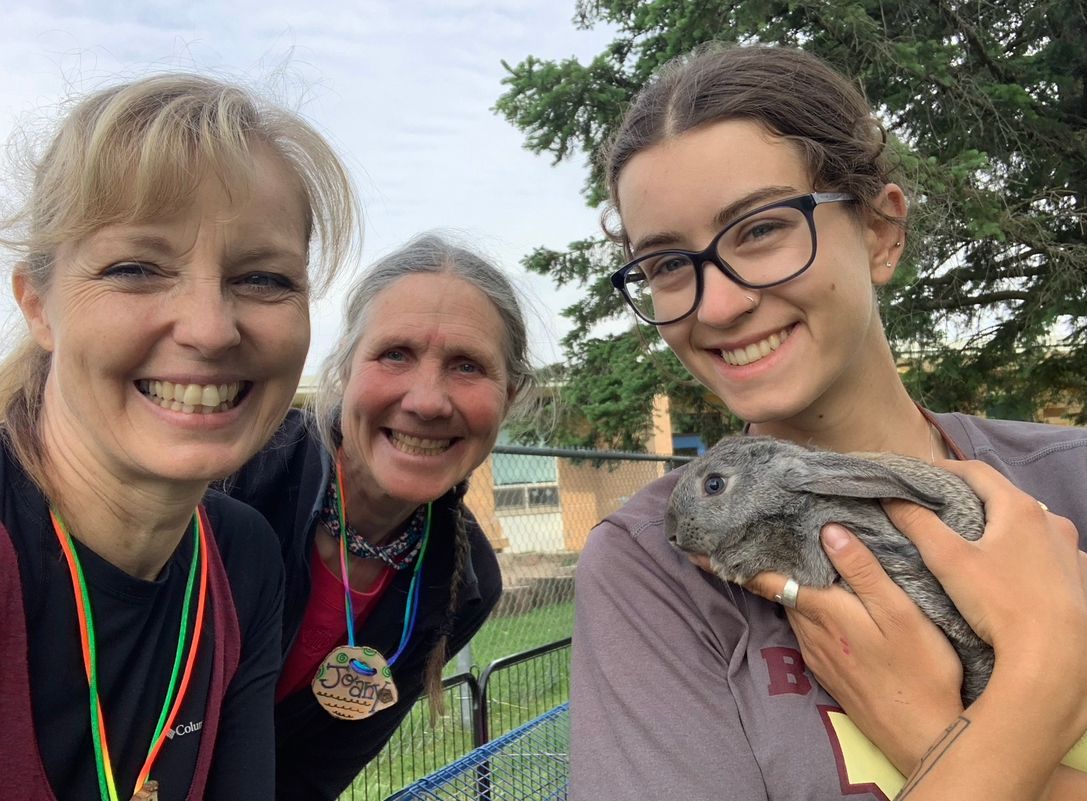 Three generations of women smiling and leaning into the frame of the image. The younger woman, about 20 years old, is holding a bunny.