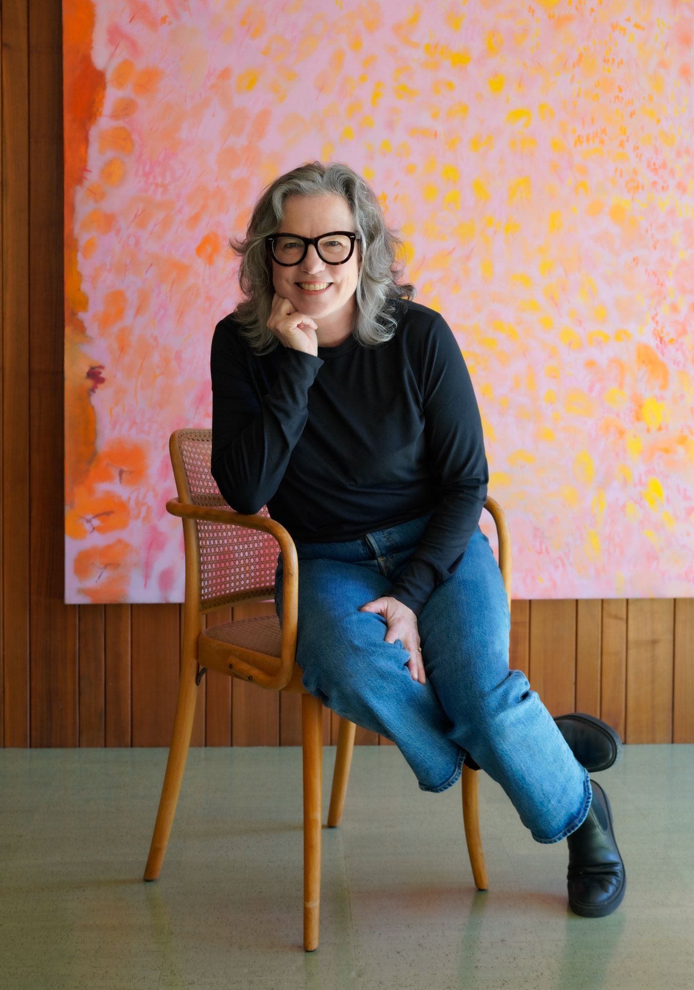A woman wearing glasses sits in a chair in front of a painting