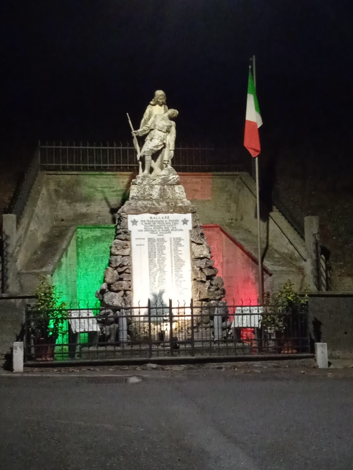 Monumento ai caduti di notte; statua raffigurante un soldato caduto, bandiera italiana, luci verdi/rosse.