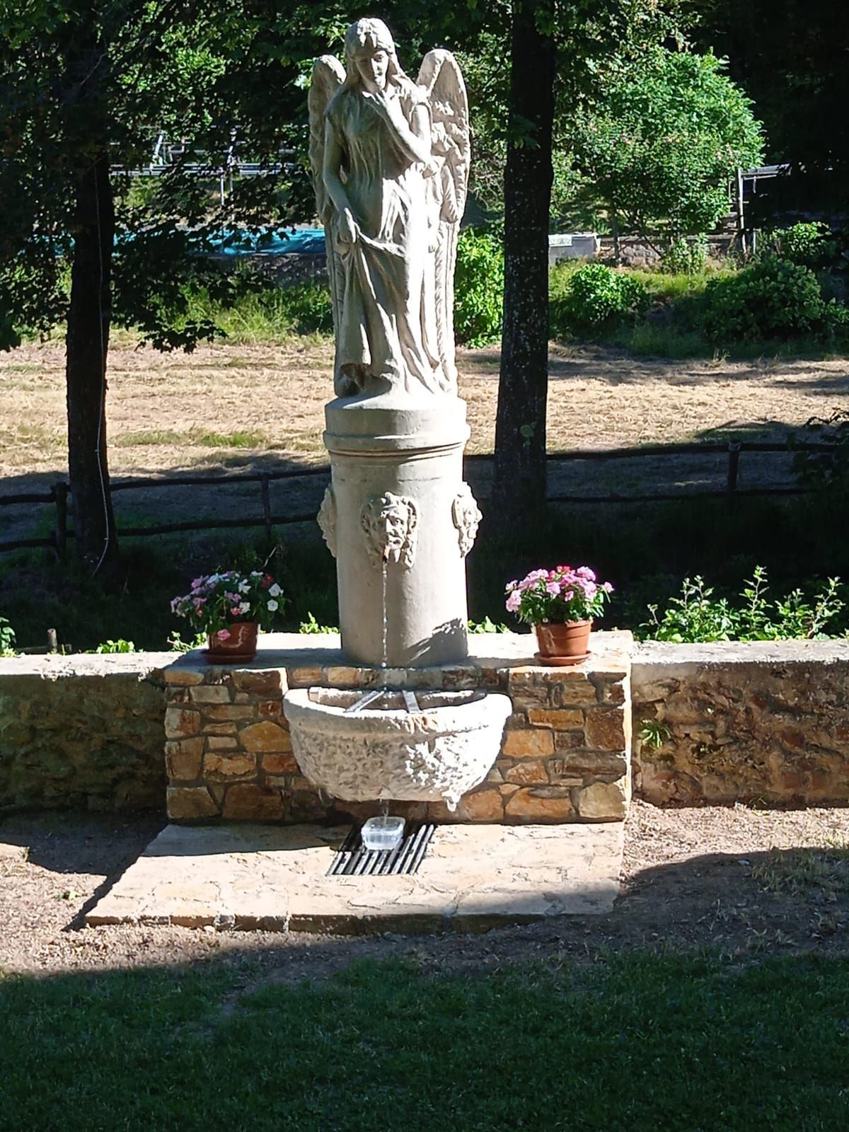 Fontana con statua di angelo in un giardino, con fiori in vaso su entrambi i lati.