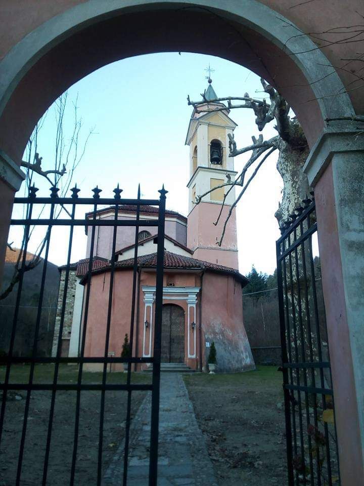 Chiesa vista attraverso un cancello in ferro battuto sotto un arco rosa, Italia. Torre con campana, pareti rosa, cielo limpido.