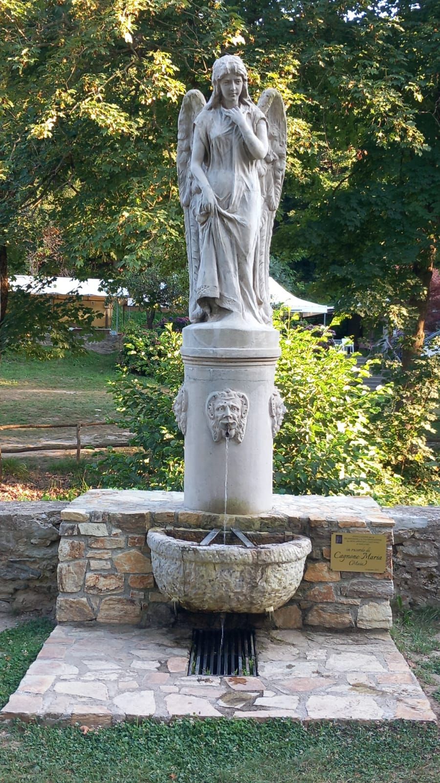 Statua di un angelo in cima a una fontana in un parco, con alberi sullo sfondo.
