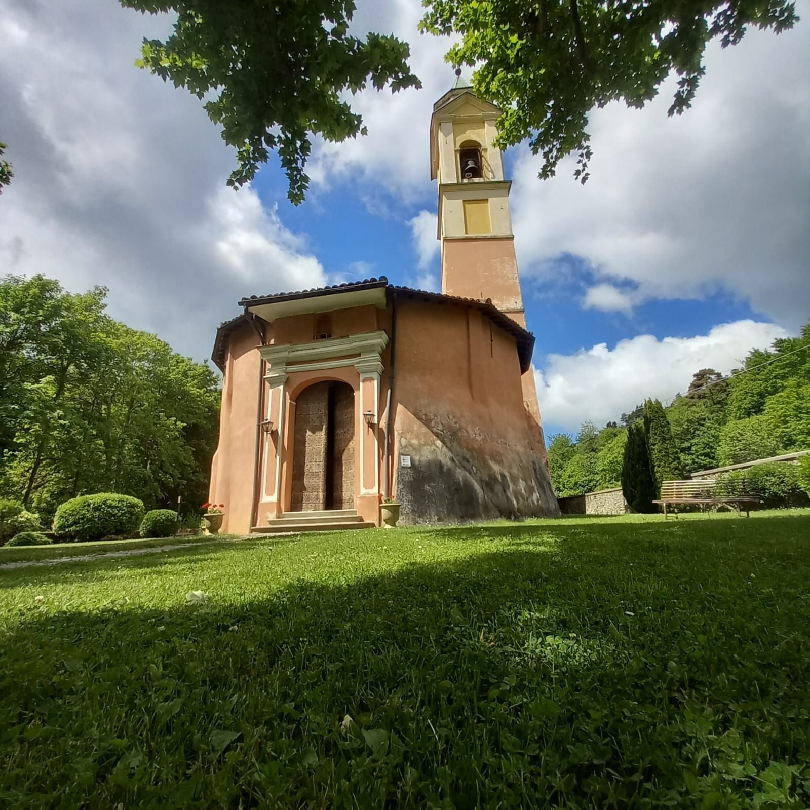 Chiesa color pesca con campanile, portale aperto, su una collina erbosa, circondata da alberi. Cielo nuvoloso.
