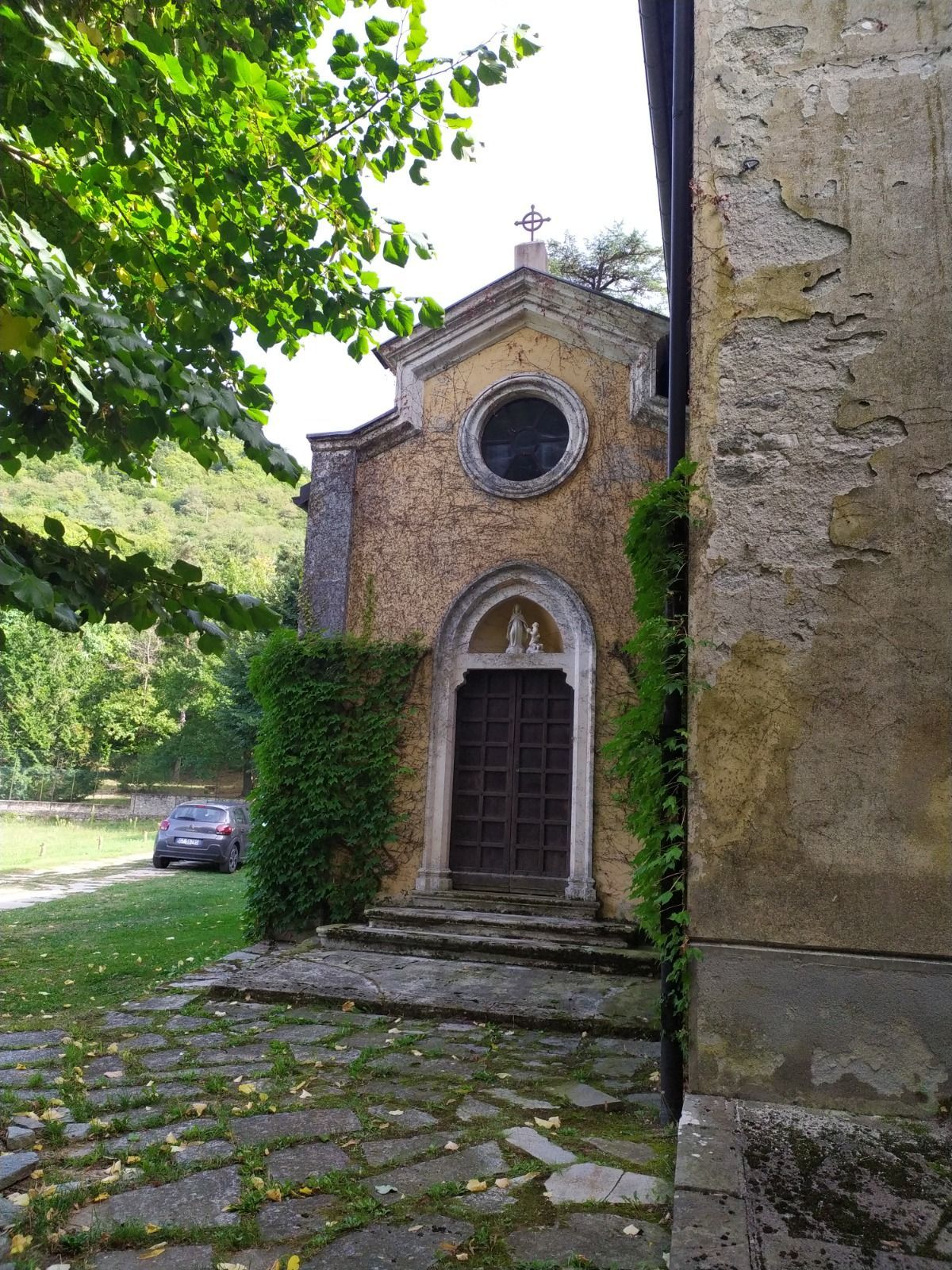 Piccola chiesa con gradini in pietra ed edera, una finestra rotonda e una porta di legno scuro.