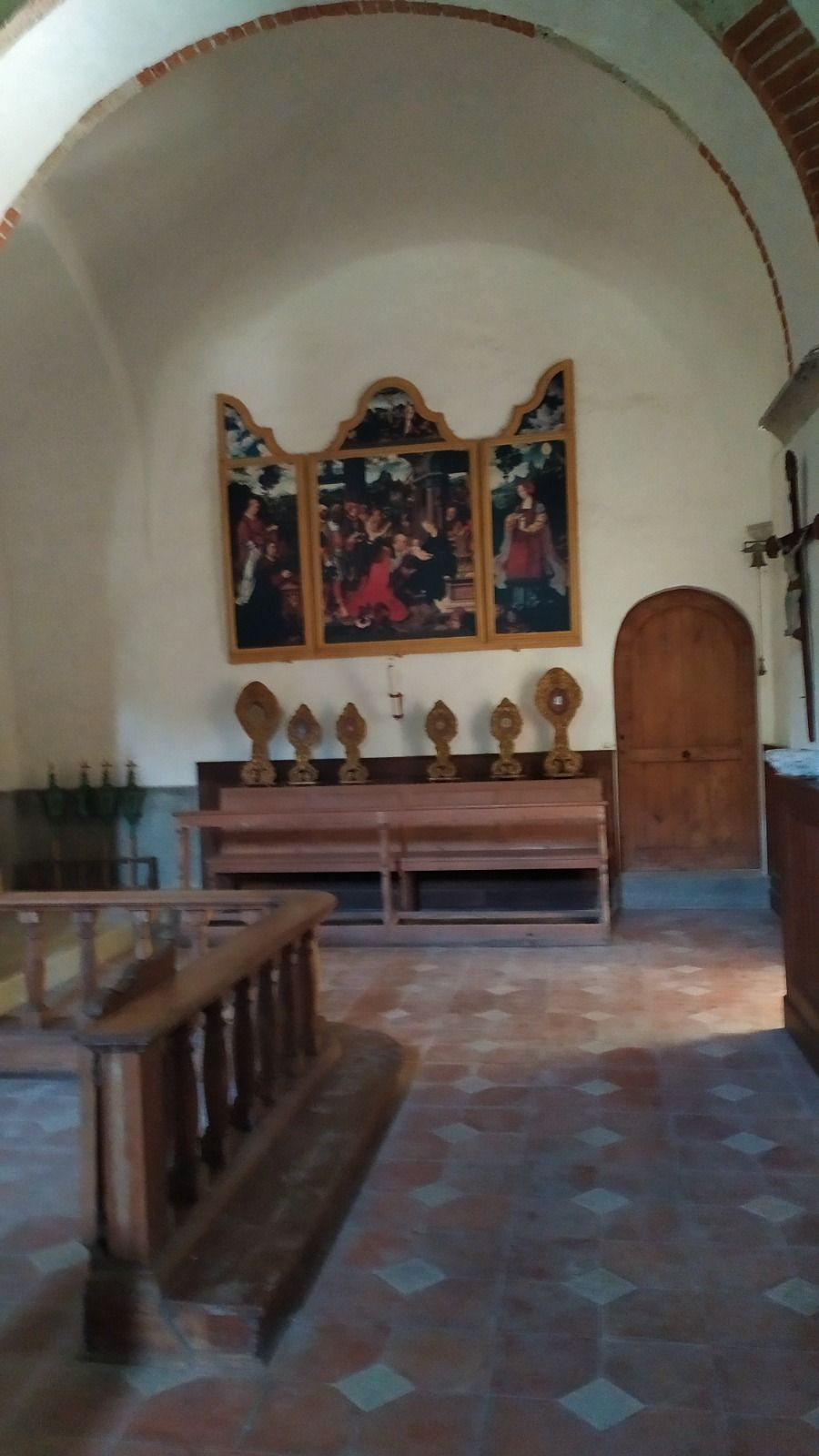 Vista interna di una cappella con opere d'arte sopra una panca, una porta di legno e un pavimento piastrellato.