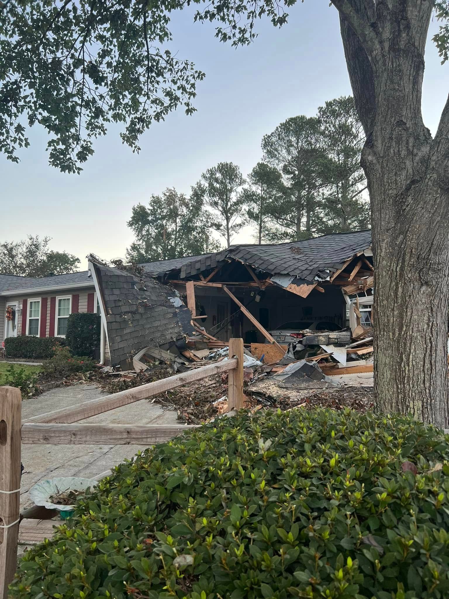 A partially collapsed building with a damaged roof, behind a wooden fence and greenery.