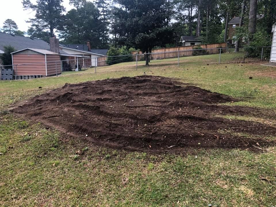 A large patch of dark brown soil in a grassy yard, ready for planting. Houses and trees are in the background.