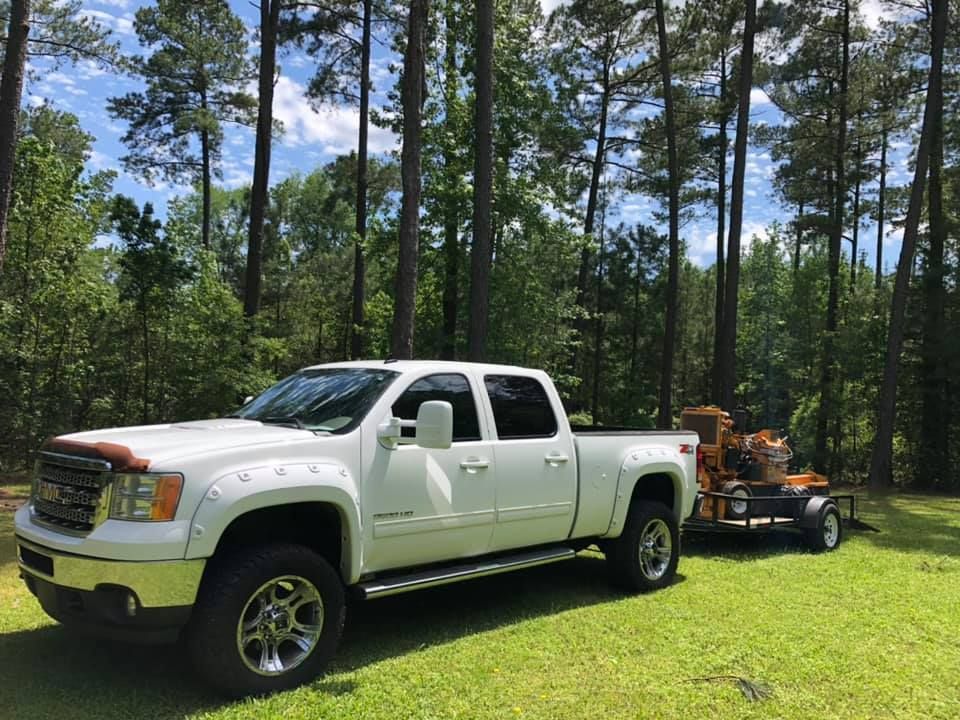 White pickup truck pulling a trailer with an orange wood chipper on green grass, trees in the background.