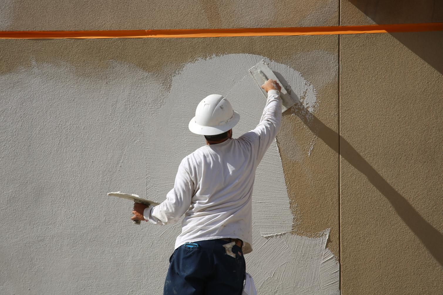 Painter in white hat applying white paint to a wall outdoors with a roller, seen from behind