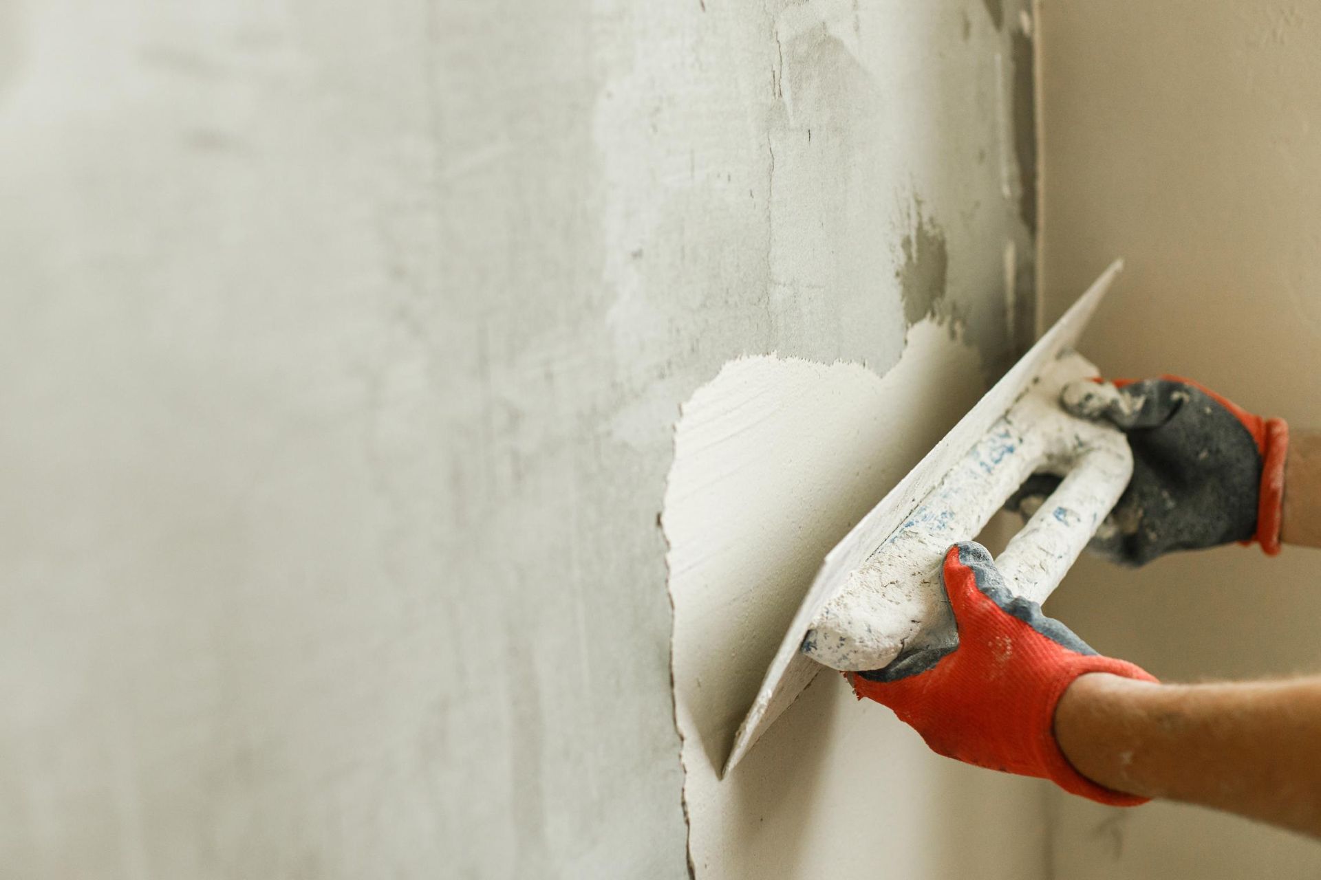 Person smoothing white plaster on a wall with a trowel, wearing red gloves.