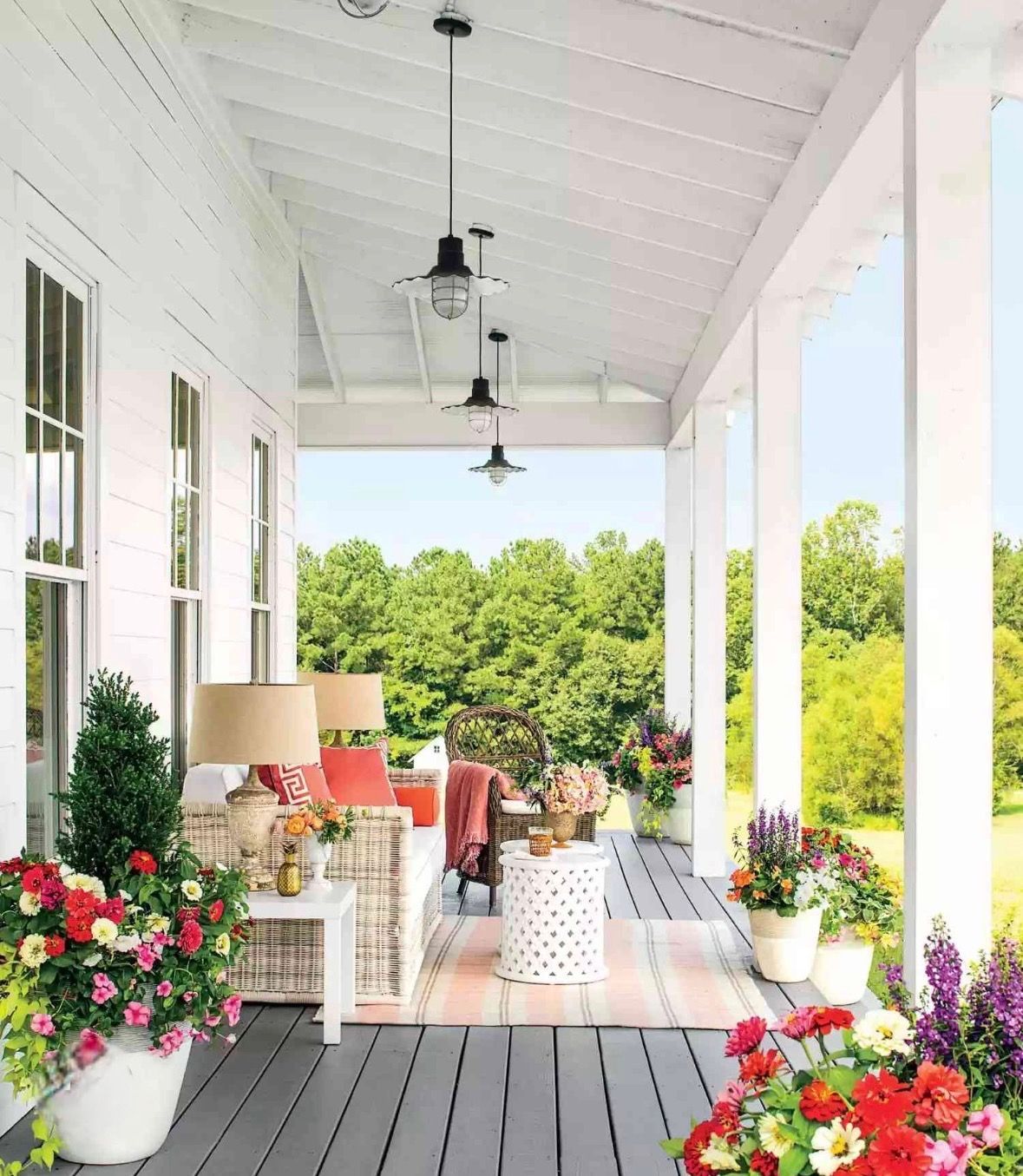 A white-painted wooden porch with grey plank flooring, furnished with cozy seating, lamps, and vibrant potted flowers.