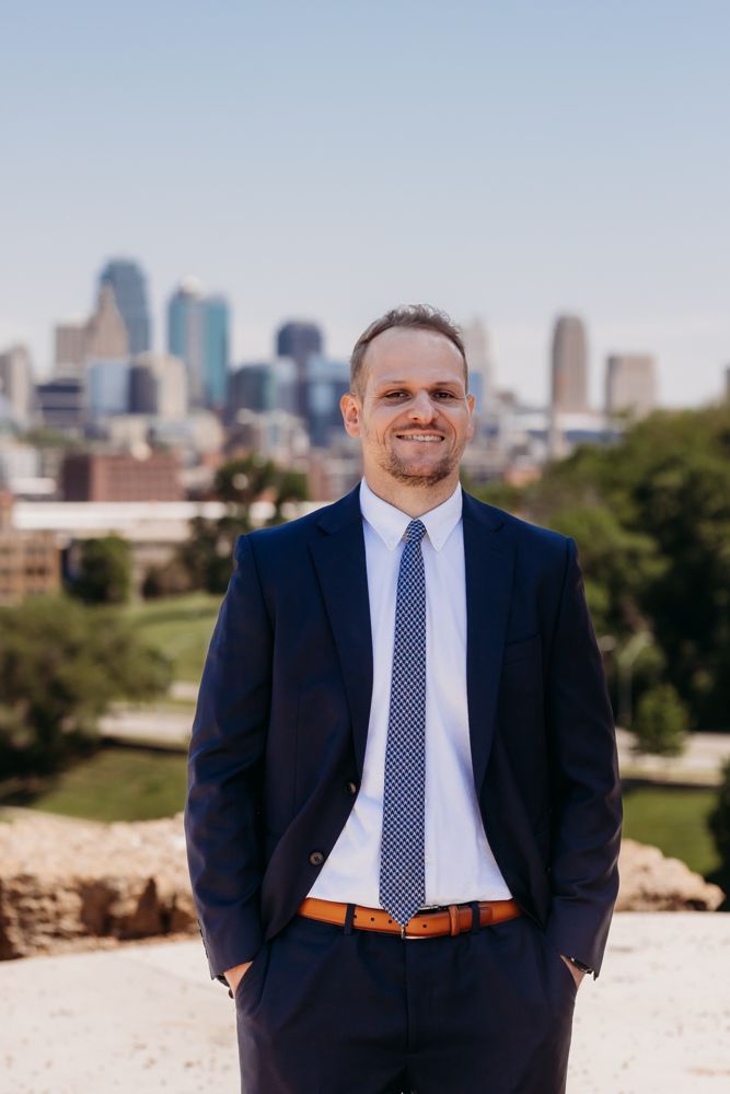 Man in navy suit and patterned tie standing outdoors with a city skyline behind him