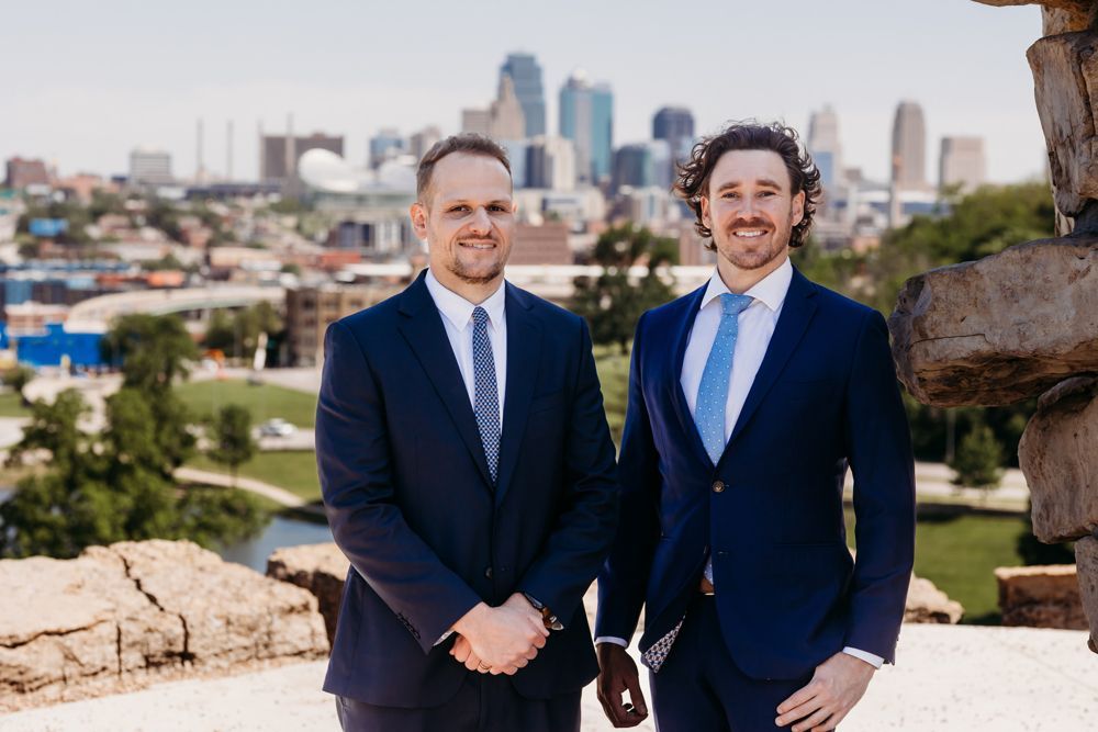 Two men in navy suits standing outdoors with a city skyline behind them