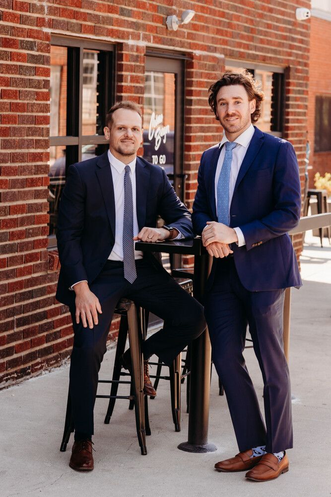 Two men in suits standing by a high table outside a brick building, holding drinks.