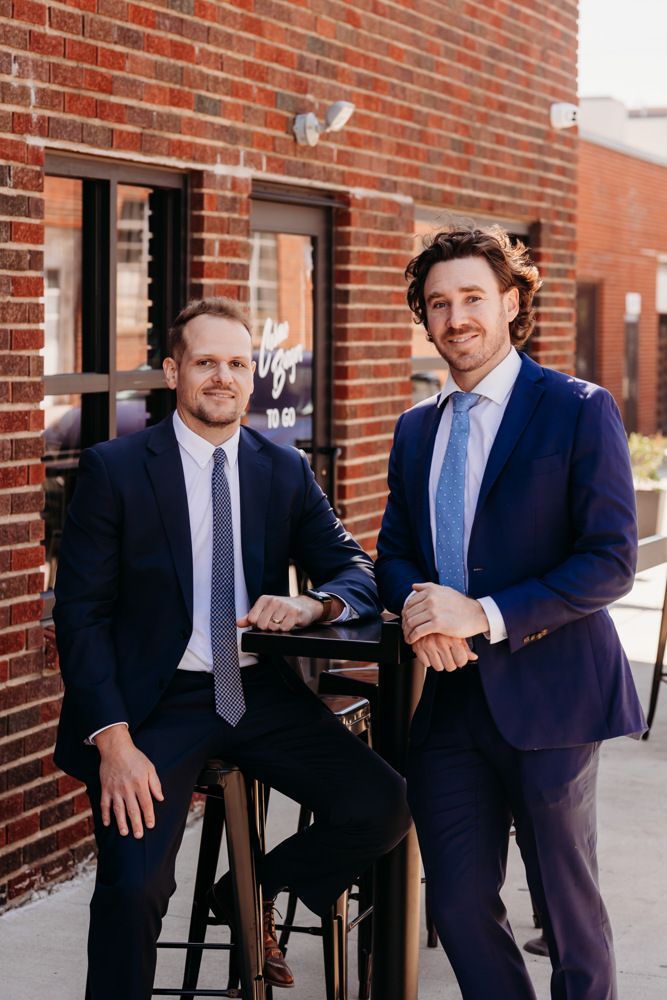 Two men in dark suits pose outside a brick building on a sunny day.