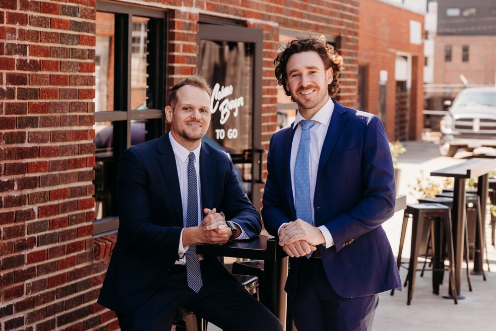Two men in suits pose outside a brick building, one seated and one standing, with outdoor tables nearby