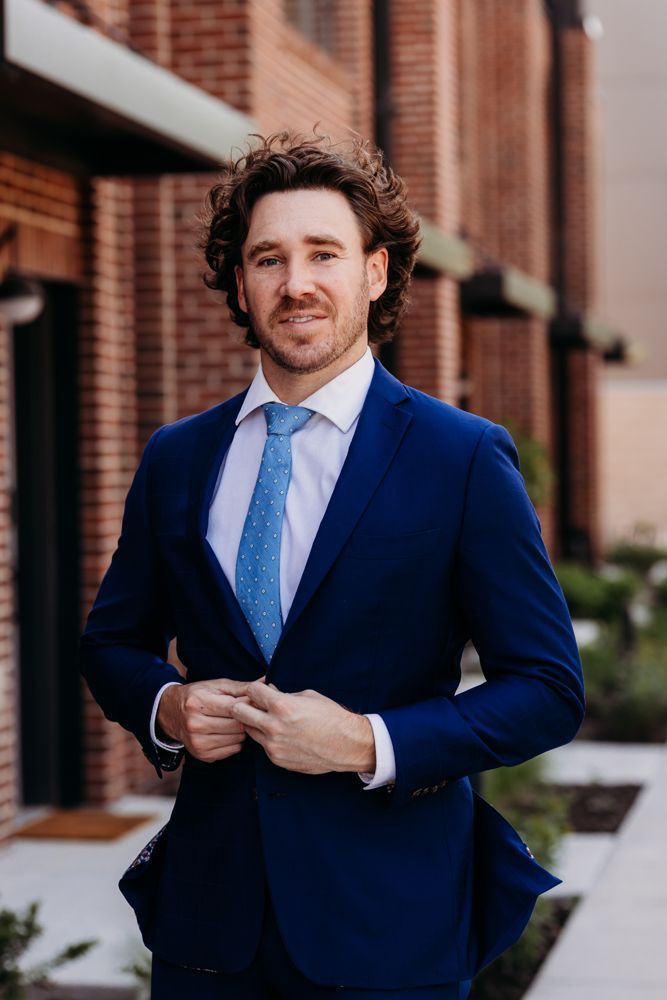 Man in a blue suit and tie standing outdoors by a brick building, adjusting his jacket.
