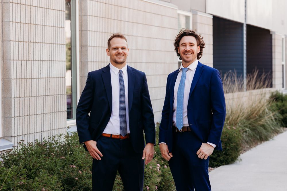 Two men in navy suits standing outdoors beside a modern building, smiling at the camera.