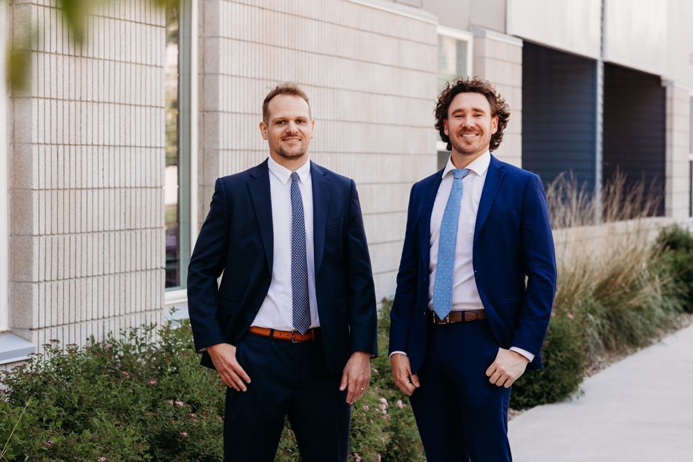 Two men in navy suits standing outside a modern building, smiling at the camera.