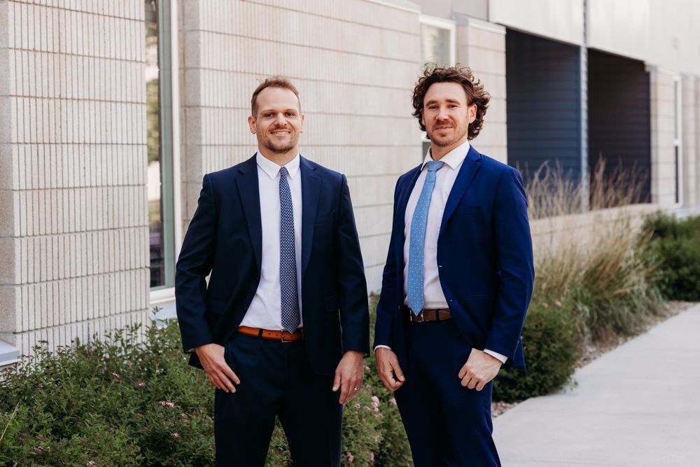 Two men in navy suits standing outdoors beside a building, smiling at the camera.