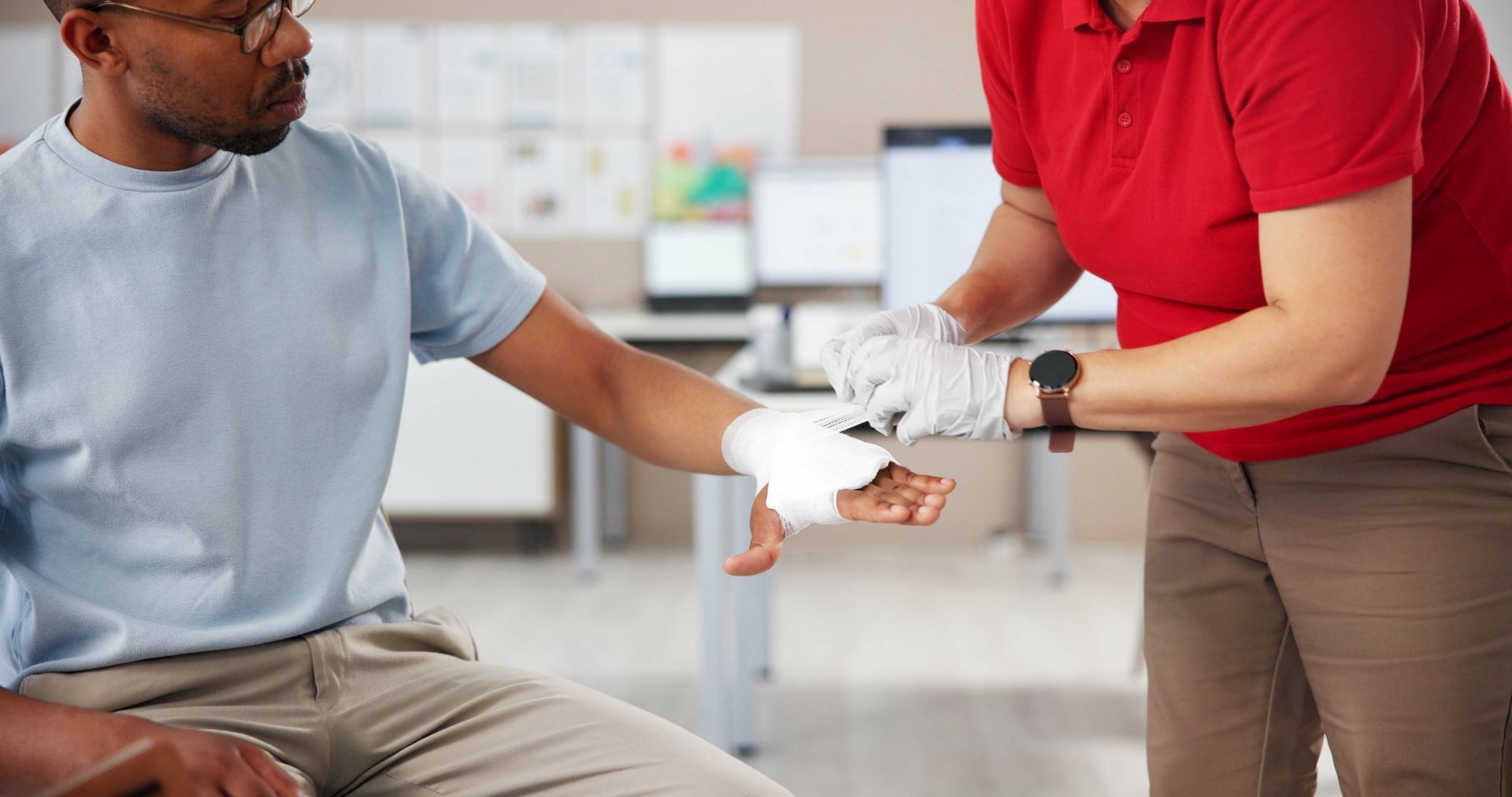 Person in a red shirt bandages a seated man’s arm in a bright clinic.
