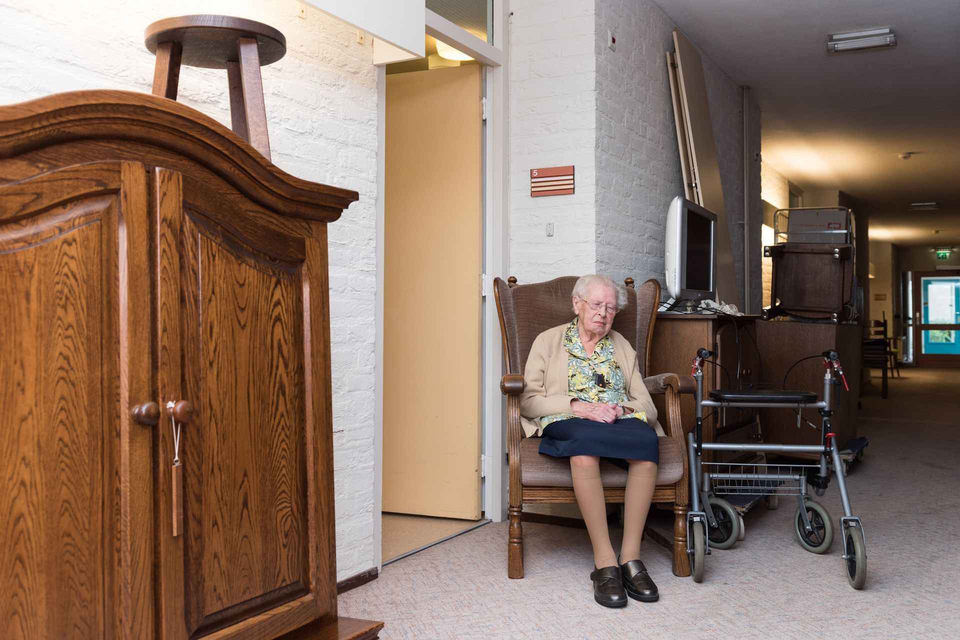 Elderly resident seated in a hallway beside a walker and wheelchair, with a wooden cabinet in the foreground