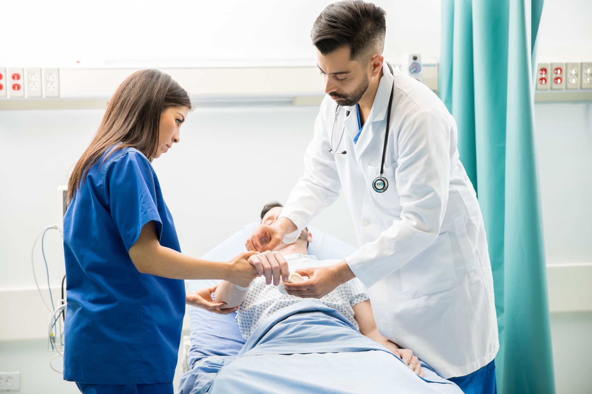 Doctor and nurse caring for a patient in a hospital bed, with a teal privacy curtain nearby.