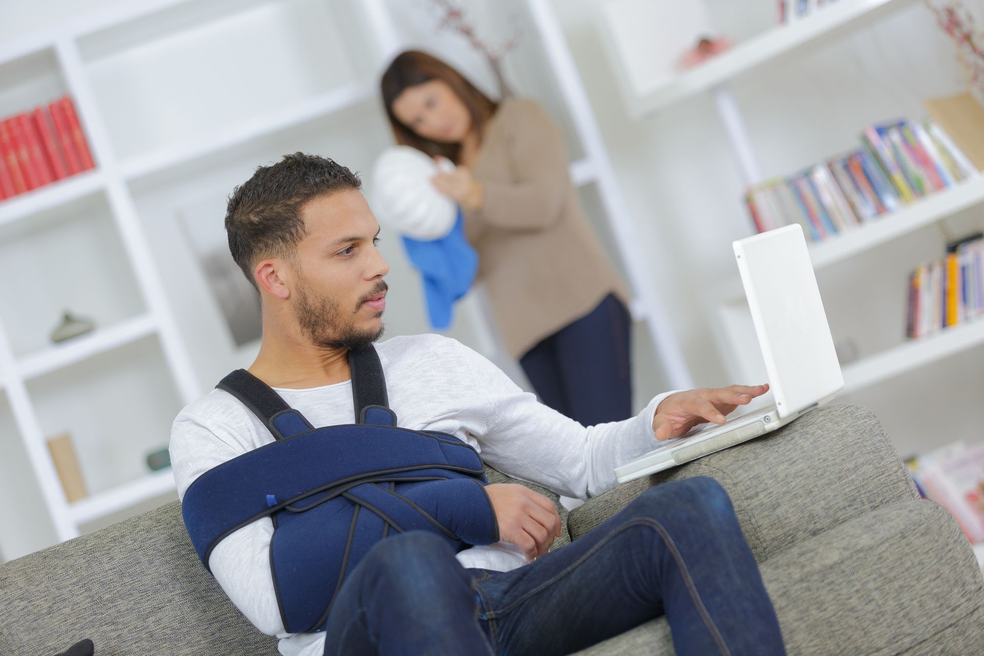 Man sitting on a couch with a laptop and arm in a sling, while a woman holds a child in the background