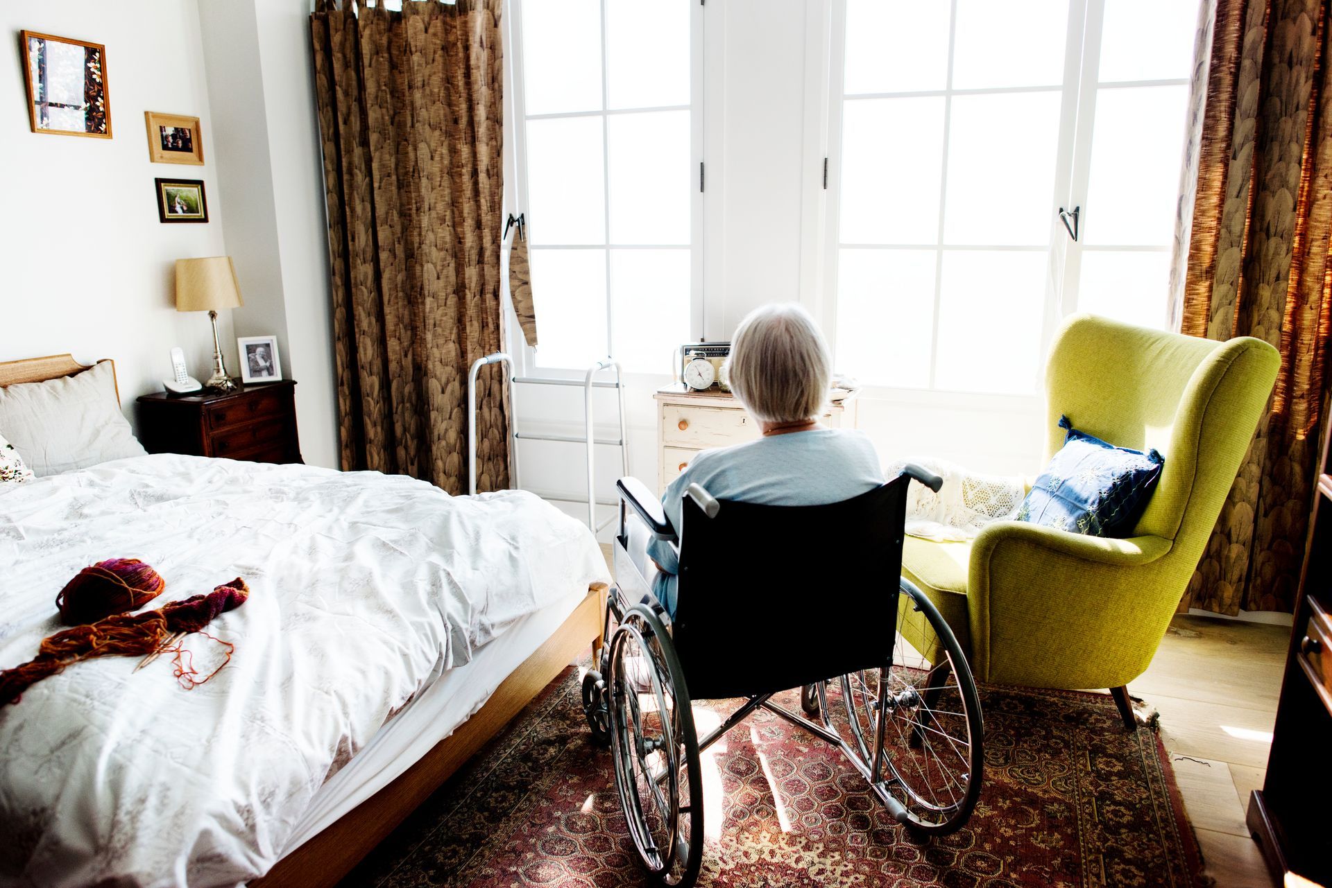 Person in wheelchair facing bright window in a cozy bedroom with bed, chair, and curtains