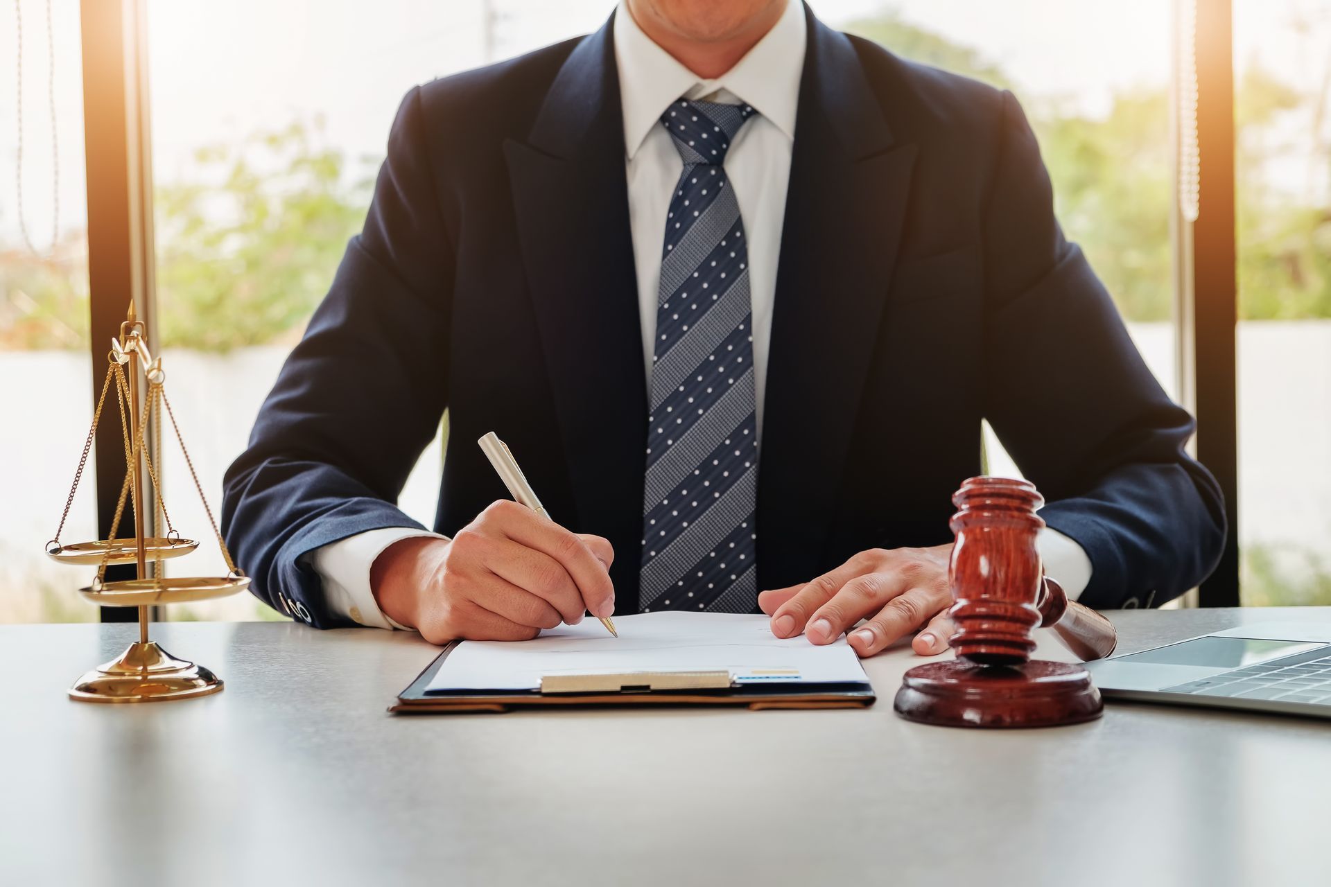 Businessperson signing documents at a desk with a gavel, scales, and a laptop in a bright office