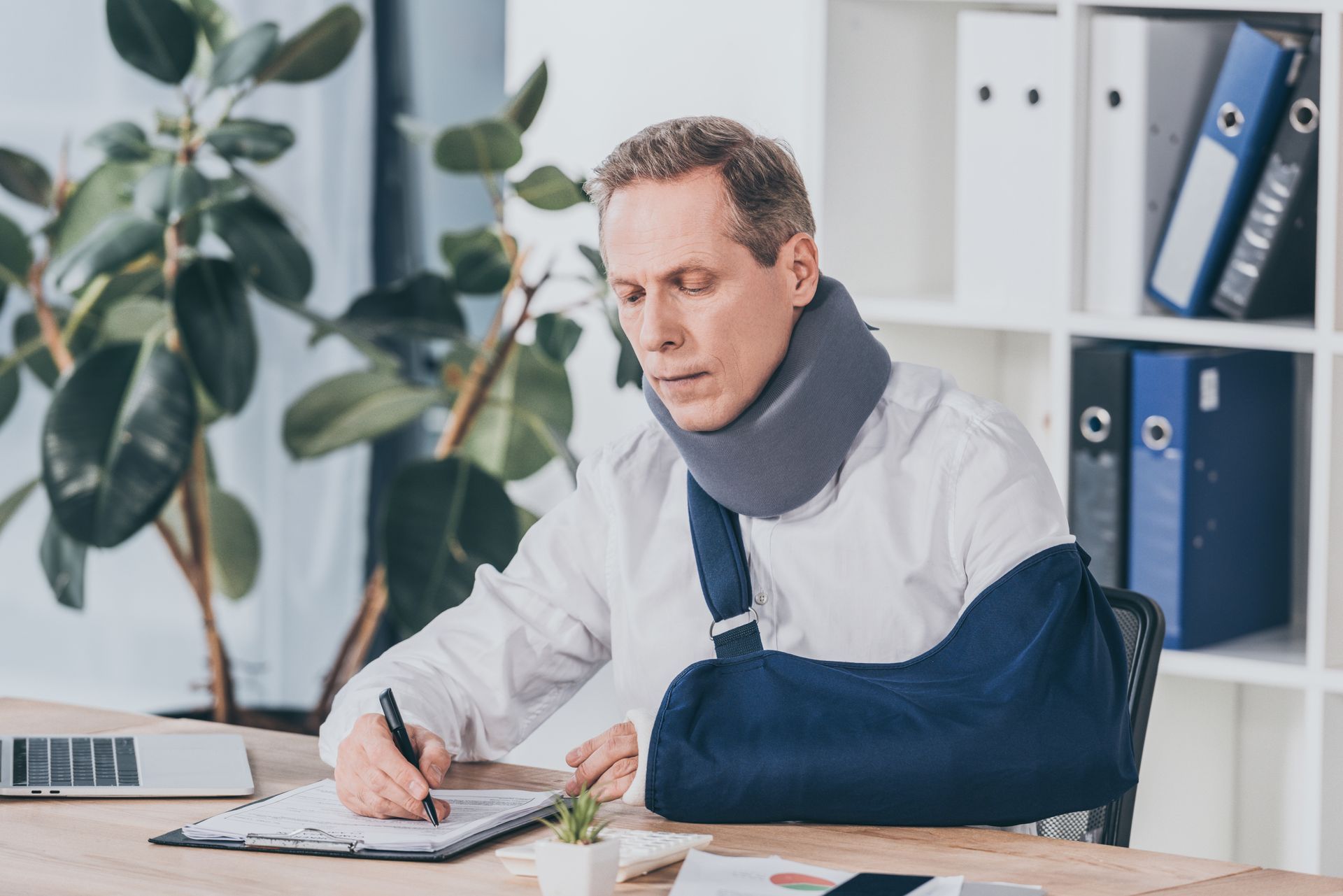 Man with arm in sling working at desk beside laptop and notebook in an office